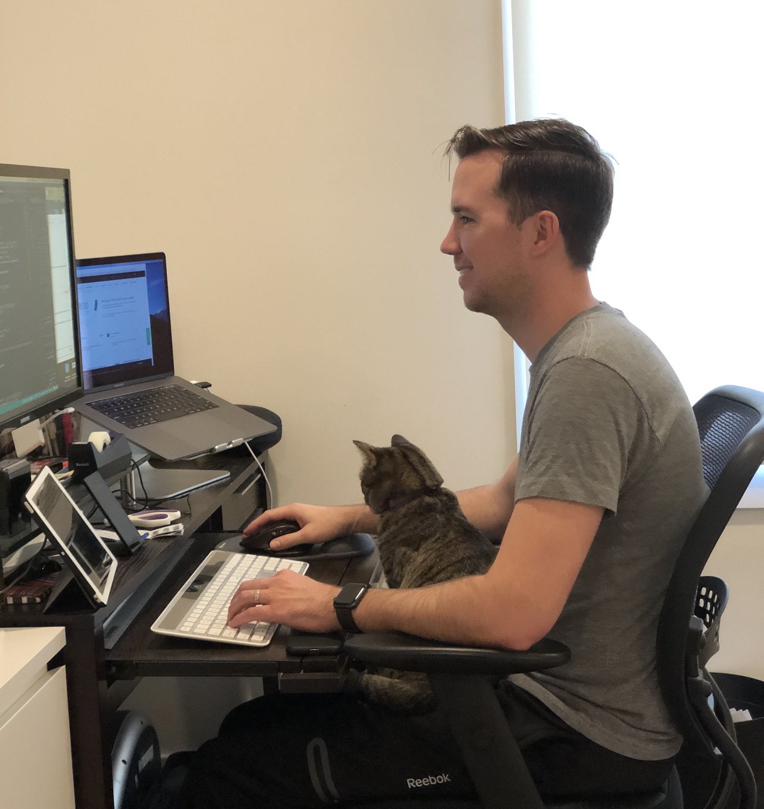 Chase McElroy at a desk with a laptop and monitor, a tabby cat sitting beside the keyboard.